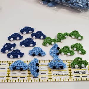 May include: A collection of small, crocheted car appliques in shades of blue and green. The cars have white wheels and black details. A ruler is in the foreground, with measurements in inches.