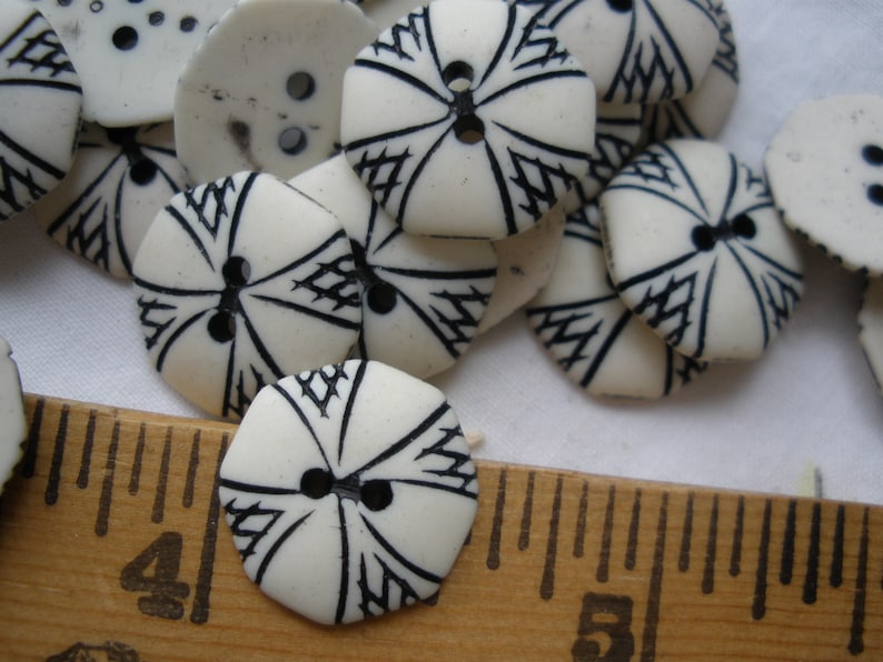 K&ouml;nnte beinhalten: Nahaufnahme eines Stapels wei&szlig;er Kn&ouml;pfe mit schwarzen geometrischen Mustern. Die Kn&ouml;pfe sind in einem zuf&auml;lligen Muster angeordnet und alle gleich gro&szlig;. Ein Holzliniall ist im unteren Teil des Bildes sichtbar.