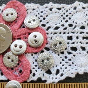 May include: A close-up of ten silver buttons with different designs. Some buttons have a simple two-hole design, while others have a more intricate design with a raised center and a decorative border. The buttons are arranged on a pink and white fabric background.