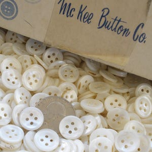 May include: A box overflowing with off-white, four-hole buttons. The buttons are round and have a pearlescent sheen. The box is labeled "Mc Kee Button Co." in blue script. A coin is visible among the buttons.