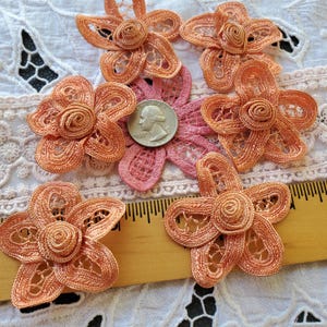 May include: Five peach-colored lace flower appliqués with a central spiral design, arranged around a silver-colored US quarter coin and a wooden ruler showing measurements in inches, placed on a white lace fabric background.