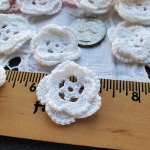 May include: White crocheted flower embellishments, approximately 1 inch in diameter, with a delicate, lacy design. The flowers are arranged on a white fabric background with a wooden ruler for scale.