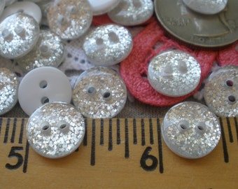 May include: A close-up of a collection of silver glitter buttons with two holes. The buttons are arranged on a pink and white fabric background with a ruler in the foreground. The ruler shows measurements in inches.