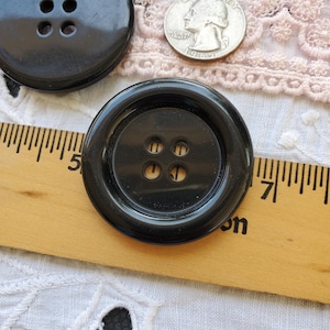May include: Close-up of two large, glossy black buttons with four holes each. A wooden ruler with inch markings and a silver quarter are also in the image. The buttons are on a white lace background.