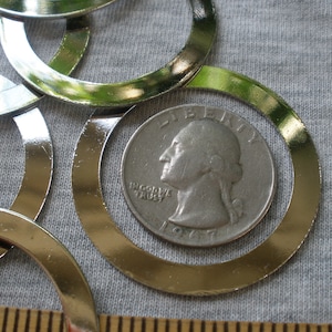 May include: A close-up of a pile of silver-colored metal rings, with one ring larger than the others. A US quarter is placed inside the larger ring for size comparison. The quarter is dated 1977 and has the inscription "IN GOD WE TRUST" visible.