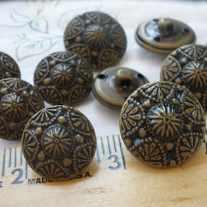 May include: A collection of antique brass-colored buttons. The buttons have a domed shape with a detailed floral and geometric pattern. Some buttons have a shank on the back for sewing. The buttons are arranged on a patterned fabric and a wooden ruler.