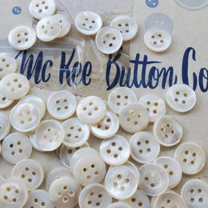 May include: A large collection of white mother of pearl buttons with four holes each. The buttons are arranged in a random pattern on a brown surface. The text "Mc Kee Button Co." is visible in the background.
