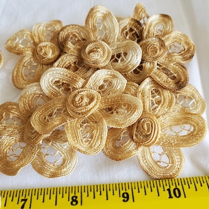 May include: Close-up of several handmade, golden-colored lace flower appliques. Each flower has a detailed, openwork design with a central spiral. The flowers are arranged on a white surface, with a yellow ruler in the foreground.
