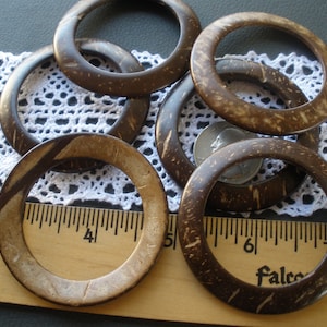 May include: Six brown coconut shell rings, some with a lighter brown color, are arranged on a white crocheted surface and a wooden ruler. The ruler is marked with inches and the text "Made in U.S.A." and "Falco".