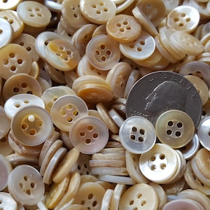 May include: A large pile of white and beige four-hole buttons. The buttons are made of a pearlescent material and are approximately the size of a US quarter.