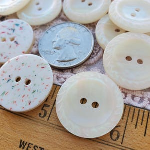 May include: A collection of round buttons in various designs. Some are white with a speckled pattern, while others are a solid pearlescent color with a decorative edge. A U.S. quarter is included for size reference. The buttons have two holes for sewing.