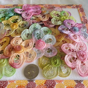 May include: A collection of colorful, handmade lace flower appliques in shades of pink, yellow, green, and orange. The delicate floral designs are arranged on a white surface with a patterned border. A US quarter is included for size reference.