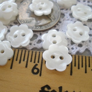 May include: A close-up of white plastic flower-shaped buttons with two holes each. The buttons are arranged on a white crocheted surface and a wooden ruler.