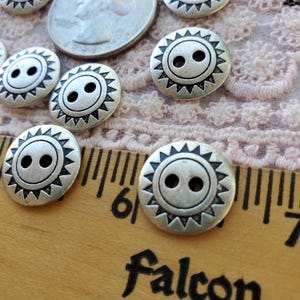 May include: Close-up of several silver-colored buttons with a sunburst design. Each button has two holes and a decorative edge. A wooden ruler with the word "Falcon" is in the foreground, along with a quarter coin and a lace fabric.