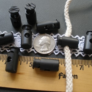 May include: Ten black plastic cord locks with a spring mechanism. The cord locks are scattered on a white lace fabric and a wooden ruler with inch markings. A quarter is included for size reference.