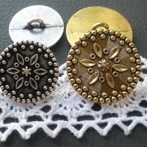 May include: Two decorative buttons with a floral design. One button is silver with a black floral design and the other is gold with a gold floral design.