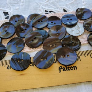 May include: A collection of 20 brown and black shell buttons with two holes each. The buttons are arranged on a white lace tablecloth with a ruler and a US quarter for scale.