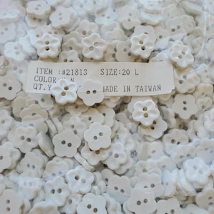 May include: A collection of white, flower-shaped buttons. The buttons have two or four holes for sewing. The image includes a label with the item number, size, color, and the text "MADE IN TAIWAN".