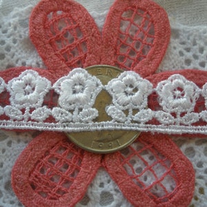 May include: White lace trim with a floral design. The trim is shown on top of a pink crocheted fabric.