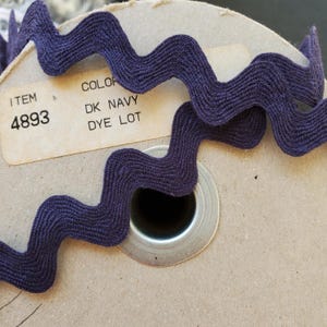 May include: Dark navy blue ric rac trim on a cardboard spool. The trim has a wavy, zig-zag design. The label reads "ITEM 4893" and "COLOR DK NAVY DYE LOT".
