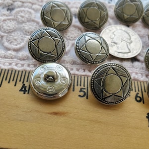 May include: A close-up of ten silver metal buttons with a star design. The buttons are arranged in a semi-circle on a wooden ruler with inch markings.
