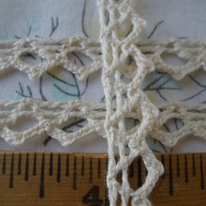 May include: Close-up of a white crocheted lace trim with a delicate pattern. The lace is shown against a ruler, indicating its size.