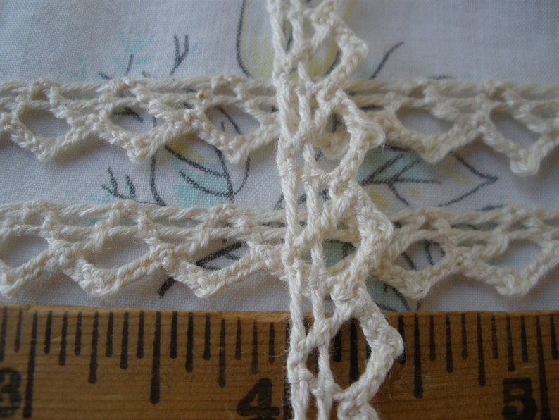 May include: Close-up of a white crocheted lace trim with a delicate pattern. The lace is shown against a ruler, indicating its size.