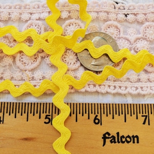 May include: Bright yellow wavy ribbon, approximately 1/2 inch wide, is displayed on a wooden ruler. A quarter coin is also present. The ribbon is against a background of pink lace.