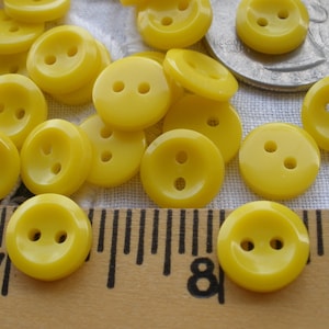 May include: A collection of bright yellow buttons with two holes each. The buttons are scattered on a white fabric surface, with a ruler and a coin in the background. The buttons are approximately 0.5 inches in diameter.