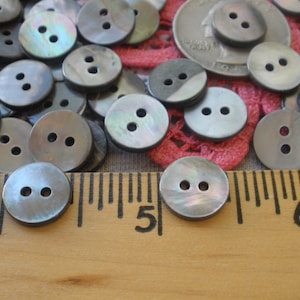 May include: A collection of round, two-hole grey iridescent shell buttons scattered near a wooden ruler. The ruler shows the buttons measure approximately 0.5 inches in diameter. A silver coin is visible among the buttons.