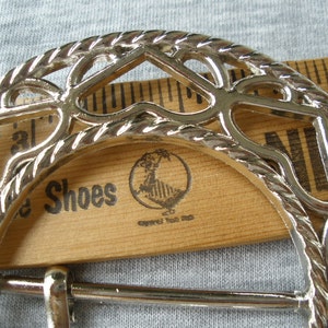 May include: Silver metal belt buckle with a decorative, openwork design. The buckle is shaped like a horseshoe and has a rope-like border. The buckle is lying on a wooden ruler with the words "Shoes" printed on it.
