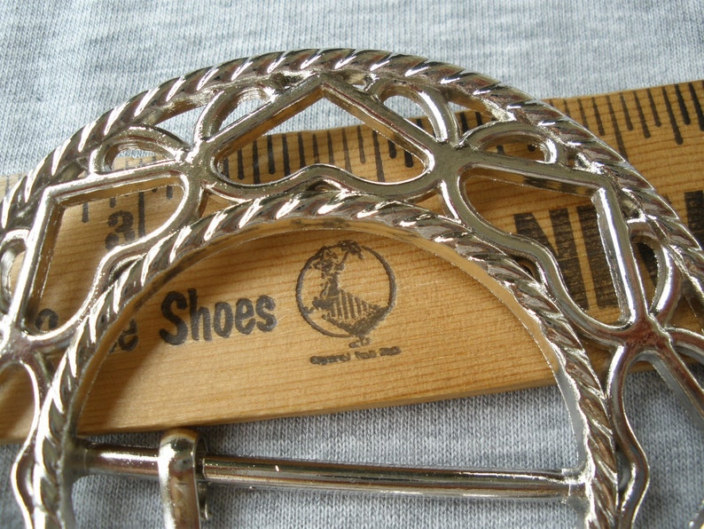 May include: Silver metal belt buckle with a decorative, openwork design. The buckle is shaped like a horseshoe and has a rope-like border. The buckle is lying on a wooden ruler with the words "Shoes" printed on it.