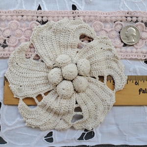 May include: Cream-colored, handmade crochet flower with a central cluster of bobbles. The flower has stylized petals and is displayed on a white surface with a pink lace trim and a ruler. A US quarter is next to the flower.