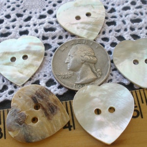 May include: Five heart-shaped buttons made of mother of pearl. The buttons have two holes each and are in various shades of white, cream, and brown.