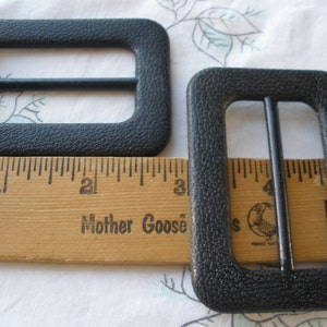 K&ouml;nnte beinhalten: Zwei schwarze Kunststoff-Schlie&szlig;en, jede mit einer rechteckigen Form und einem einzelnen Metallb&uuml;gel. Die Schlie&szlig;en sind etwa 2,5 cm breit und 3,8 cm lang. Die Schlie&szlig;en werden neben einem Holzlineal mit dem aufgedruckten Text "Mother Goose" gezeigt.