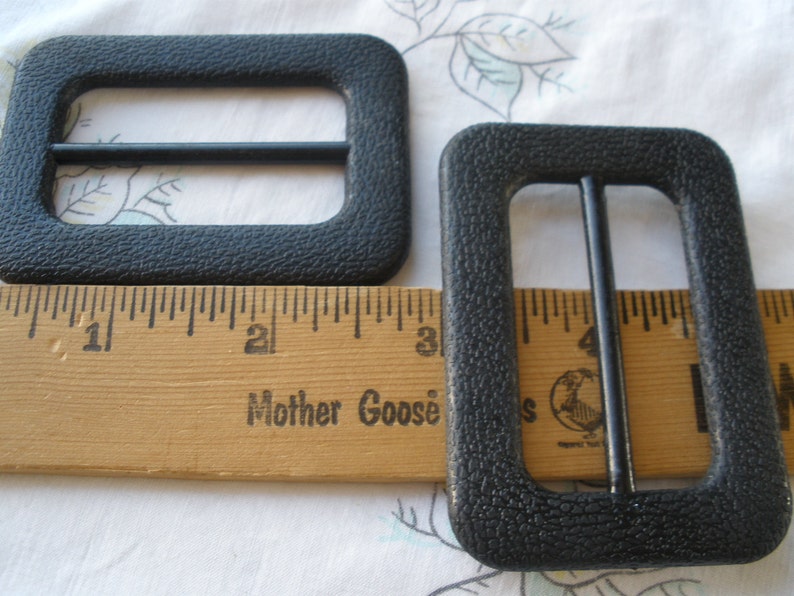 K&ouml;nnte beinhalten: Zwei schwarze Kunststoff-Schlie&szlig;en, jede mit einer rechteckigen Form und einem einzelnen Metallb&uuml;gel. Die Schlie&szlig;en sind etwa 2,5 cm breit und 3,8 cm lang. Die Schlie&szlig;en werden neben einem Holzlineal mit dem aufgedruckten Text "Mother Goose" gezeigt.