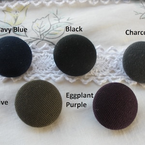 May include: Six fabric covered buttons in various colors: navy blue, black, charcoal, olive, eggplant purple, and dark purple. The buttons are arranged in a row on a white lace background.