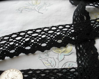 Puede incluir: Borde de encaje de crochet negro con un delicado patrón calado. El encaje se muestra sobre un fondo de tela blanca con un diseño floral.