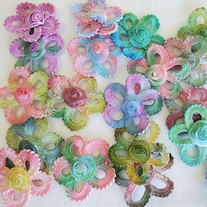 May include: Assortment of crocheted flower embellishments in various shades of pink, blue, and green. Each flower has a center swirl and multiple layers of delicate crocheted petals.