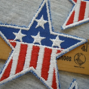 May include: Two red, white, and blue star-shaped patches with a blue background and white stars. The patches are made of fabric and have a stitched edge.