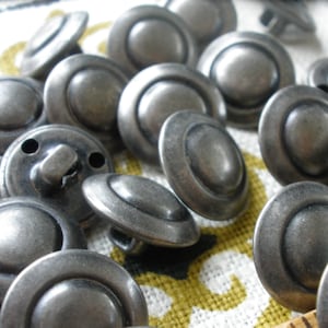 May include: A collection of silver-toned metal buttons with a raised center and a slightly rounded edge. The buttons are arranged in a random pattern on a fabric surface.