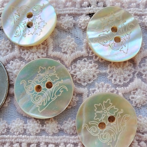 May include: Close-up of several iridescent mother-of-pearl buttons. Each button features a floral design with two holes for sewing. The buttons have a pearlescent sheen and are set against a pink lace background.