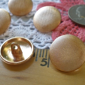 May include: A close-up of ten gold-colored buttons with a textured surface. The buttons are round and have a shank on the back. The buttons are arranged in a random pattern on a white lace surface.