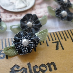 May include: Close-up of several small, handmade fabric flowers with a denim-like texture and a pearl bead center. Each flower is attached to a light green ribbon. The flowers are arranged on a wooden ruler.