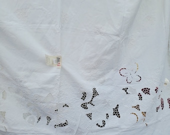 May include: White tablecloth with cut-out floral and butterfly designs. The fabric has a textured appearance with embossed floral patterns. The cut-out details create a decorative border along the edge of the cloth. A tag is attached to the tablecloth.