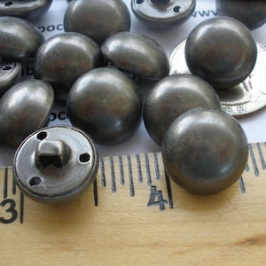 May include: A close-up of a group of 14 silver metal buttons. The buttons are round and have a smooth, shiny finish. They are arranged on a wooden ruler with markings in inches.