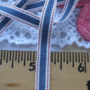 May include: Close-up of a blue, white, and red striped ribbon. The ribbon is approximately 0.5 inches wide and is lying on a wooden ruler. The ribbon has a woven texture and is likely used for crafting or sewing projects.
