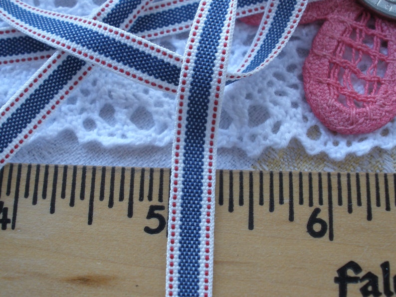 May include: Close-up of a blue, white, and red striped ribbon. The ribbon is approximately 0.5 inches wide and is lying on a wooden ruler. The ribbon has a woven texture and is likely used for crafting or sewing projects.