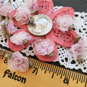 May include: A collection of 10 small, lavender-colored fabric rose embellishments with a satin ribbon bow. The roses are arranged on a pink and white crocheted doily.