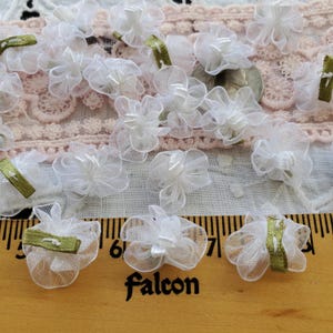May include: A collection of white tulle flower embellishments with green ribbon accents. The flowers are arranged on a white fabric background with a wooden ruler in the foreground.